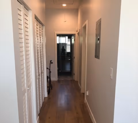A narrow hallway with wooden flooring and white walls. On the left side, there are white louvered closet doors, and on the right side, there is a wall with a small electrical panel. At the end of the hallway, there is a doorway leading to another room with a glass door.