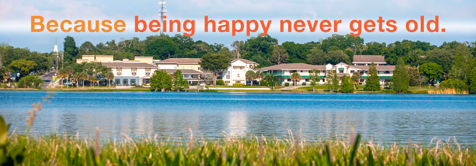 View of Westminster Lakeland senior living facility across a calm lake with green grass and trees in the foreground and background. The sky is clear and blue, and the text 'Because being happy never gets old.' is overlaid at the top in orange and red gradient.