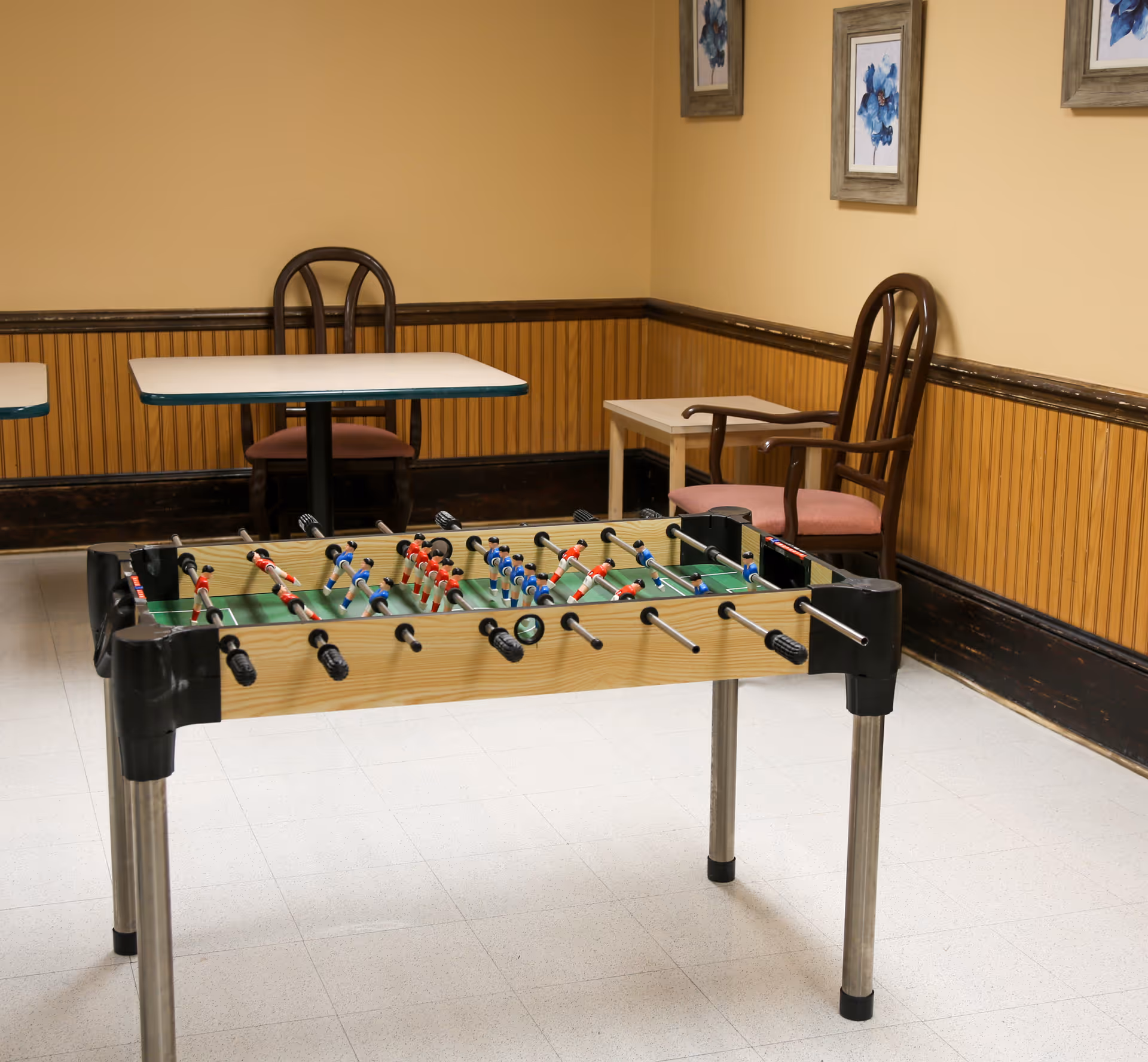 A room with a foosball table in the foreground and two tables with chairs against the walls. The walls are painted yellow with wooden paneling on the lower half, and two framed floral pictures hang on the wall.