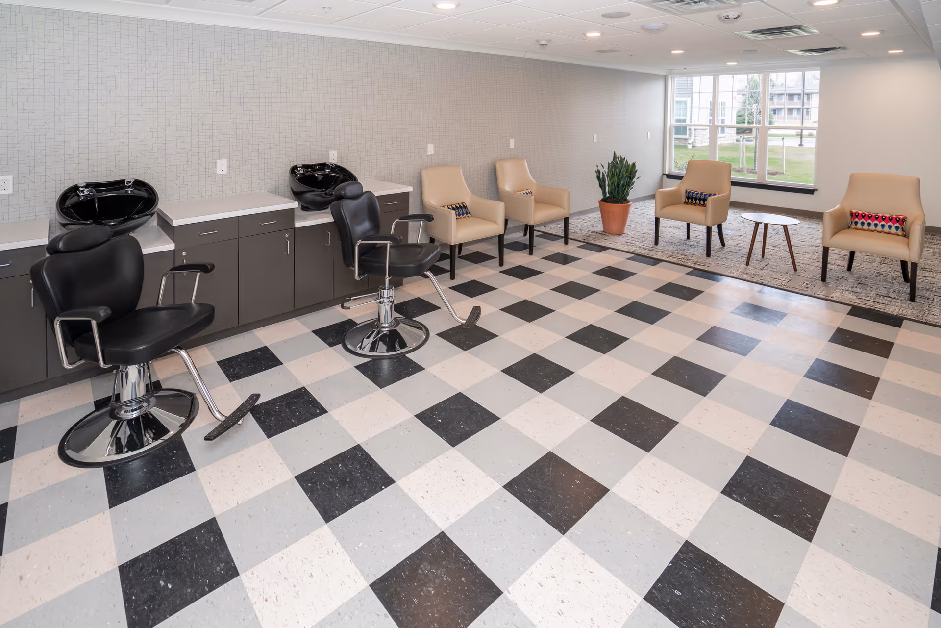 Interior view of a salon area in a senior living facility with two black salon chairs in front of black wash basins and a row of beige armchairs with colorful pillows near a large window, all set on a black, white, and gray checkered floor.