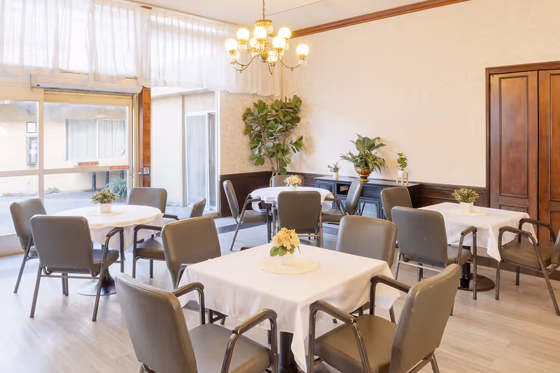 A bright dining room with several square and round tables covered with white tablecloths, each adorned with small floral centerpieces. The room has gray chairs around the tables, large windows letting in natural light, a chandelier hanging from the ceiling, and potted plants along the walls.