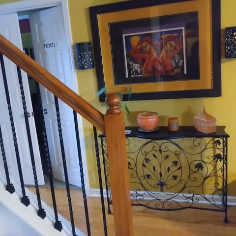Interior view of a hallway with a wooden staircase railing on the left, a decorative black metal console table with a floral design against a yellow wall, topped with various decorative items including a pink pot and a small box. Above the table hangs a framed colorful painting of two women. A white door labeled 'PRIVATE' is partially open in the background.