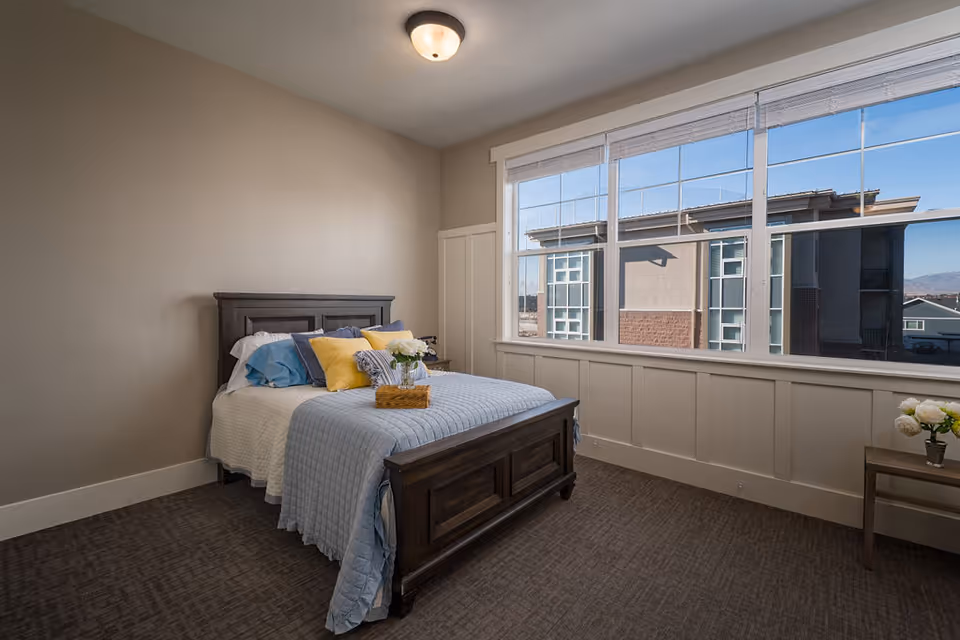 Bedroom with a wooden bed dressed in blue bedding and yellow pillows and a large window overlooking adjacent buildings.
