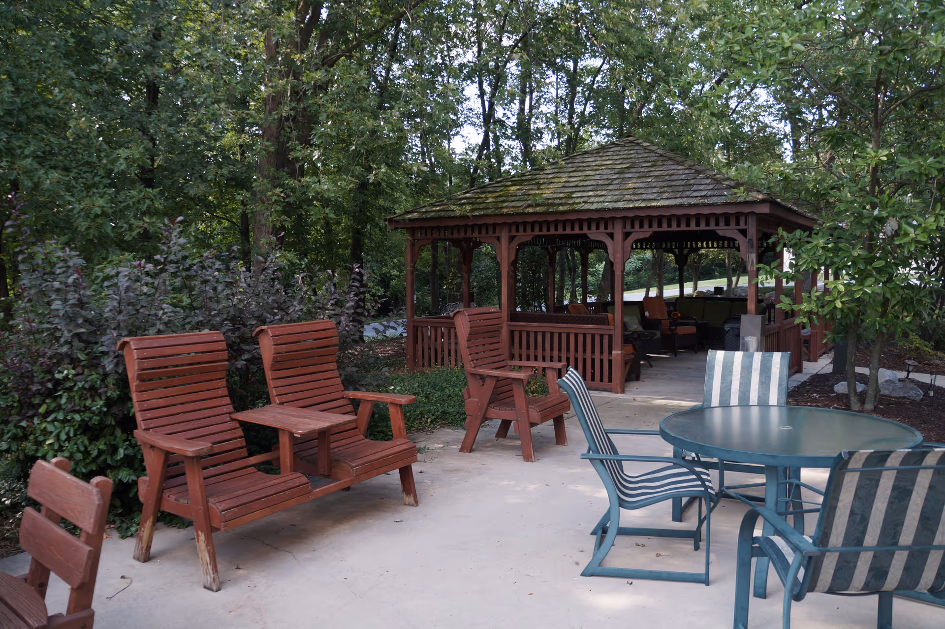 Patio with wooden bench-style chairs, a round table with striped chairs, and a covered gazebo surrounded by trees.