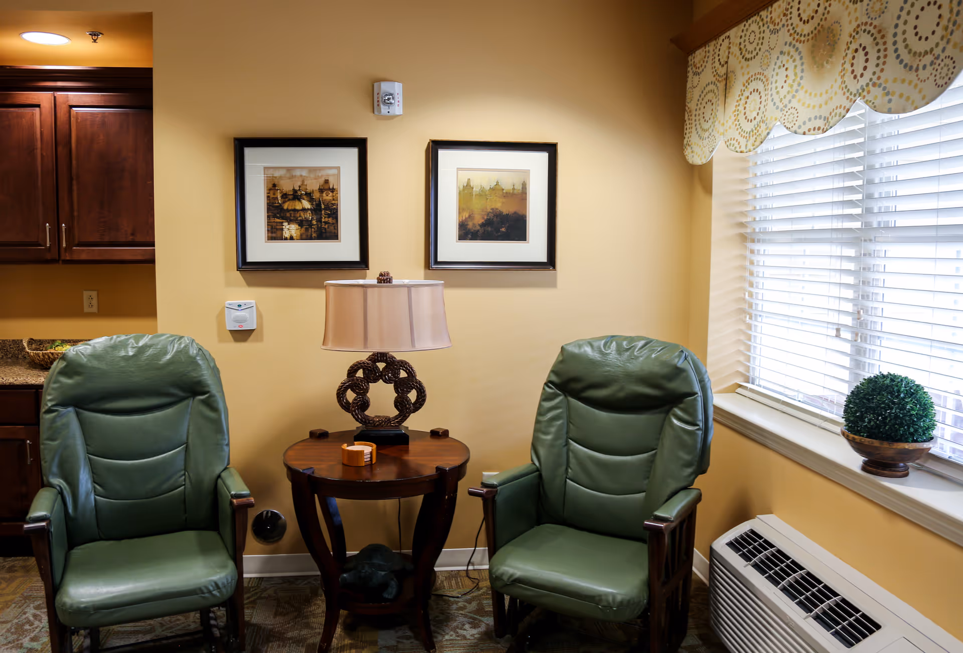 A cozy interior corner of a senior living facility with two green cushioned armchairs placed on either side of a round wooden side table with a decorative lamp. The wall behind has two framed pictures, and there is a window with white blinds and a patterned valance. A small potted plant sits on the windowsill above an air conditioning unit.