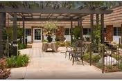 Sunlit courtyard with a pergola, table and chairs, hanging plants, and doors leading into the building.