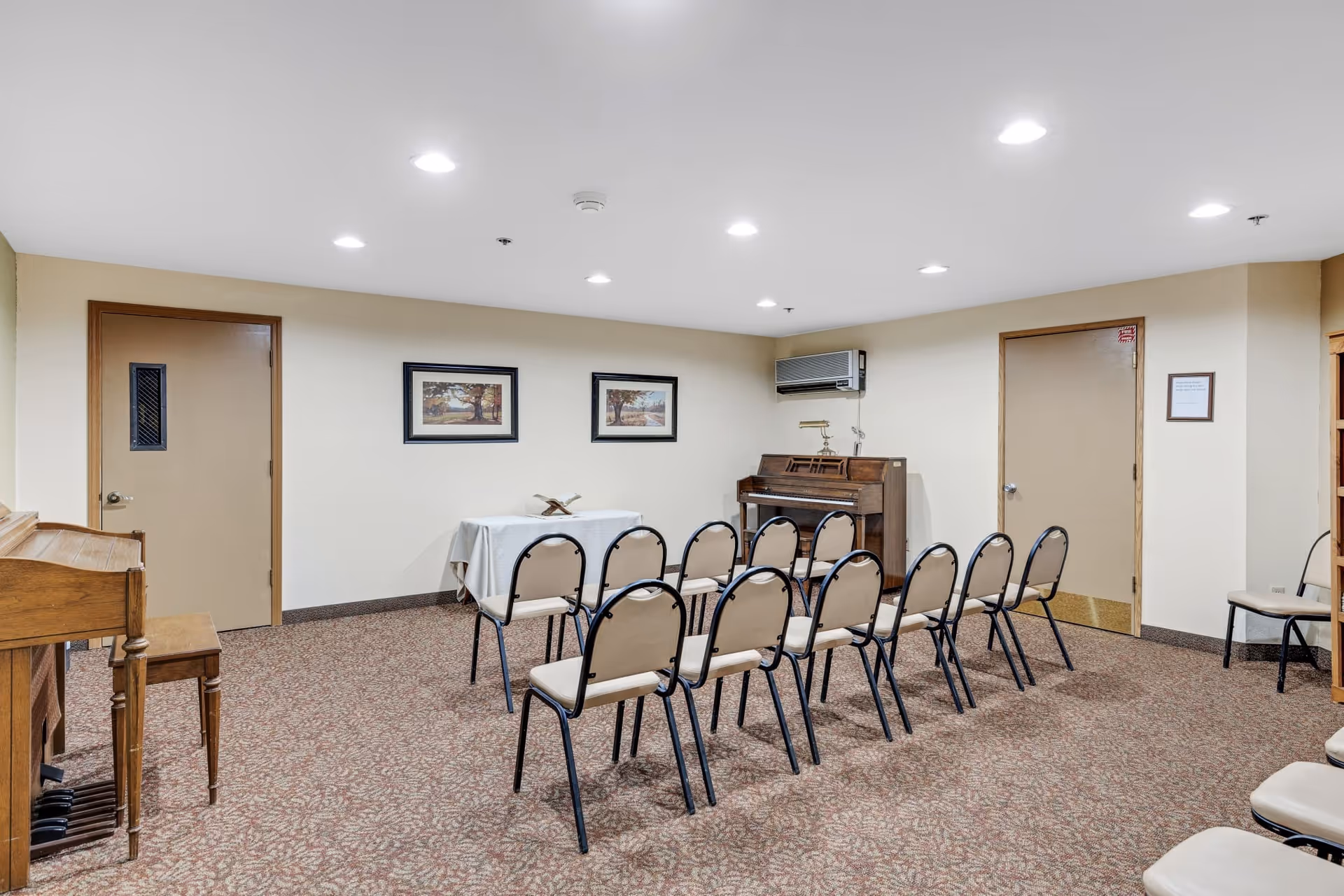 A small meeting or activity room with two rows of beige chairs facing a table covered with a white cloth. There is a piano against the wall and two framed pictures hanging above the table. The room has beige walls, a patterned carpet, and two closed doors.