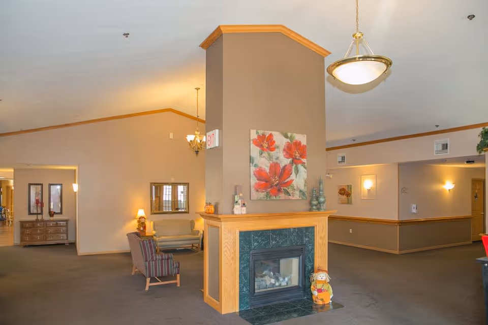 Interior view of a senior living facility common area with a central fireplace surrounded by green tiles and wooden trim. Above the fireplace is a painting of red flowers. There are chairs and a small sofa arranged near the fireplace, a wooden dresser with framed pictures on the left, and warm lighting fixtures on the walls and ceiling. A small decorative scarecrow figure is placed on the floor next to the fireplace.