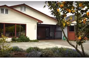 Exterior view of a single-story building with white siding and a red roof trim, featuring large windows and a gated entrance. There is a concrete driveway in front and a tree with orange fruit on the right side.