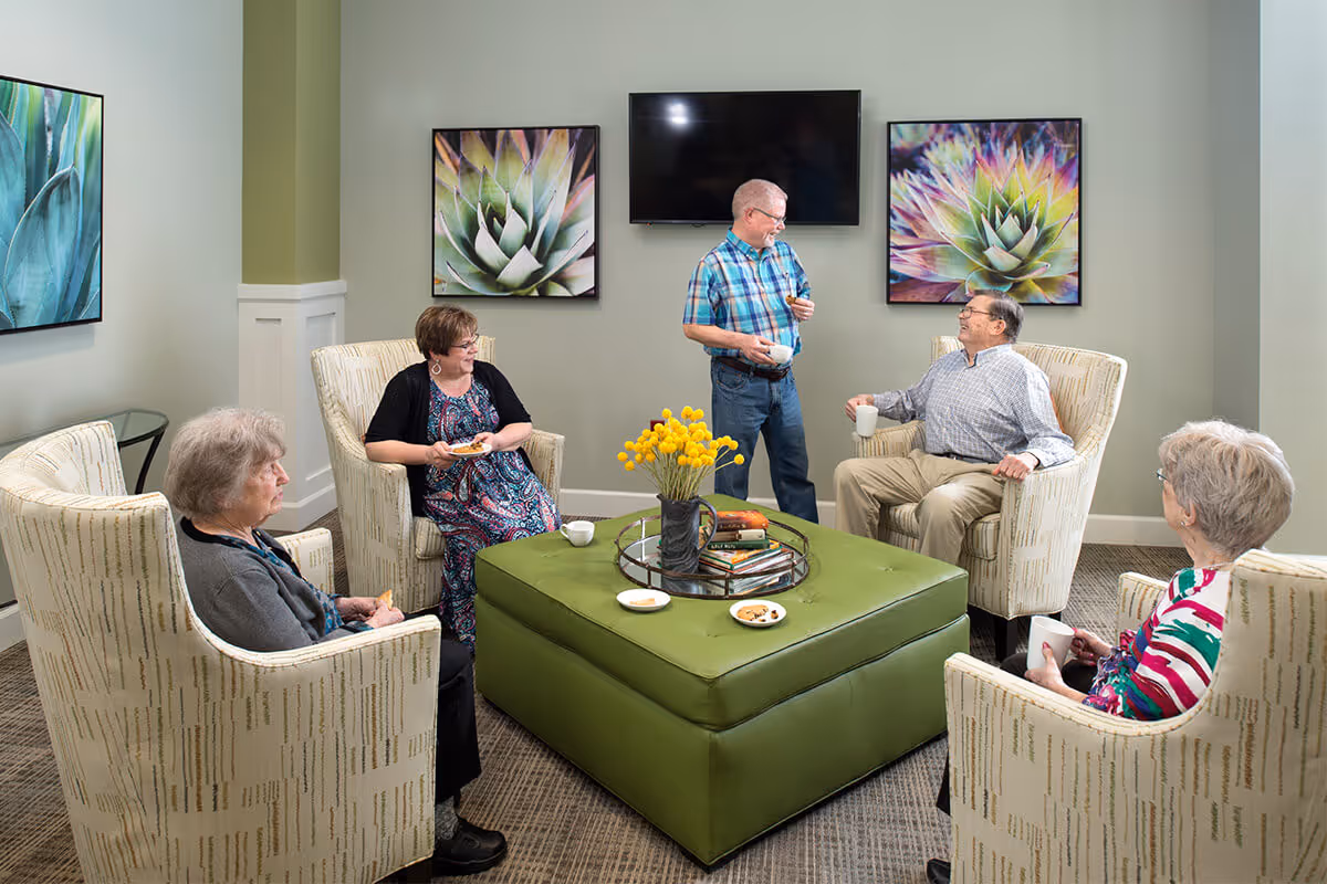 A group of five elderly people sitting and standing in a cozy living room area with four armchairs arranged around a large green ottoman. They are engaged in conversation, holding cups and plates with snacks. The room has light-colored walls decorated with three large framed succulent plant artworks and a flat-screen TV mounted on the wall.