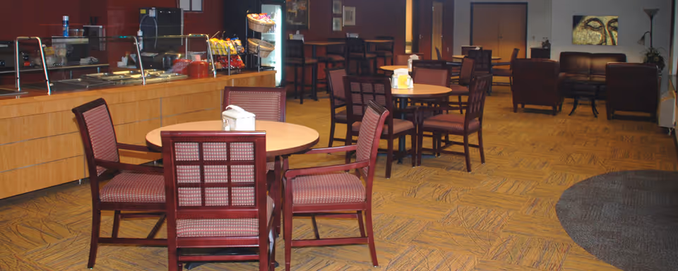 Dining area with round tables and wooden chairs, a buffet counter on the left, and lounge seating in the background.