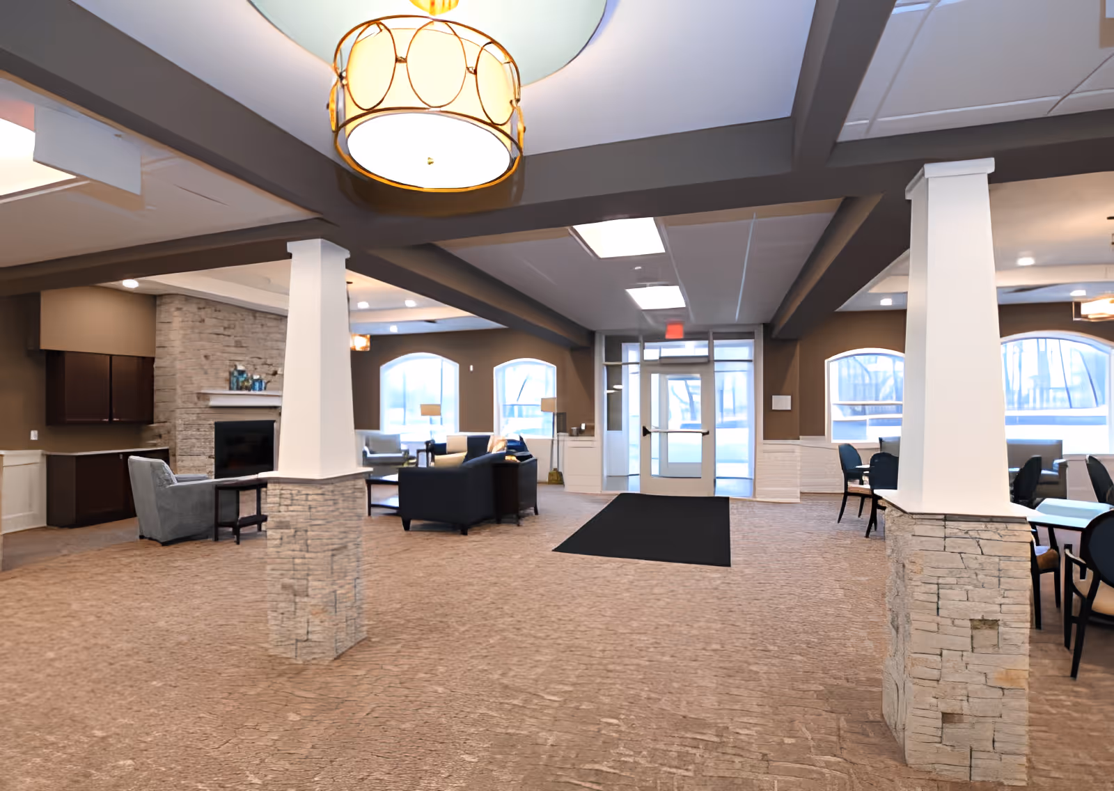 Spacious assisted living facility common area with beige carpet, stone pillars, and brown walls. The room features a seating area with sofas and armchairs near a stone fireplace, large windows letting in natural light, and a dining area with tables and chairs. Ceiling lights and a large hanging lamp illuminate the space, and a glass door leads outside.