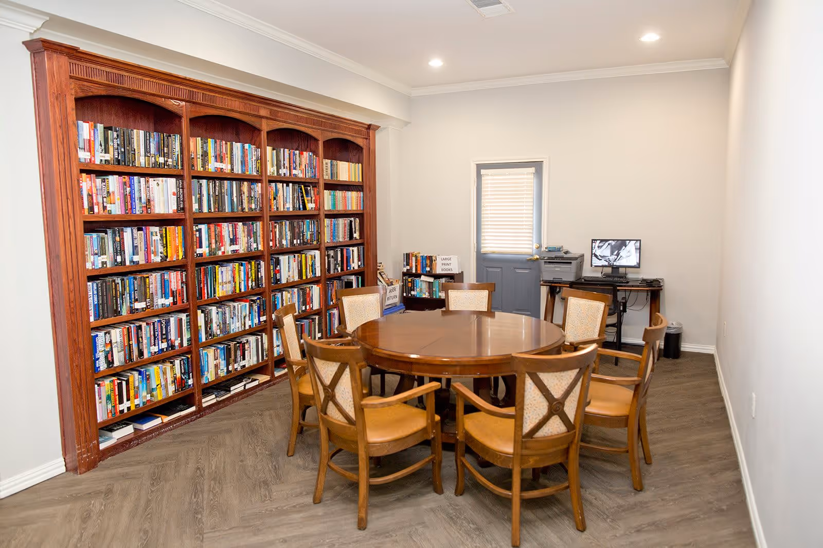 A cozy room with a large wooden bookshelf filled with books along one wall, a round wooden table surrounded by six wooden chairs with cushioned seats and backs, and a small desk with a computer and printer near a door with a window covered by blinds.