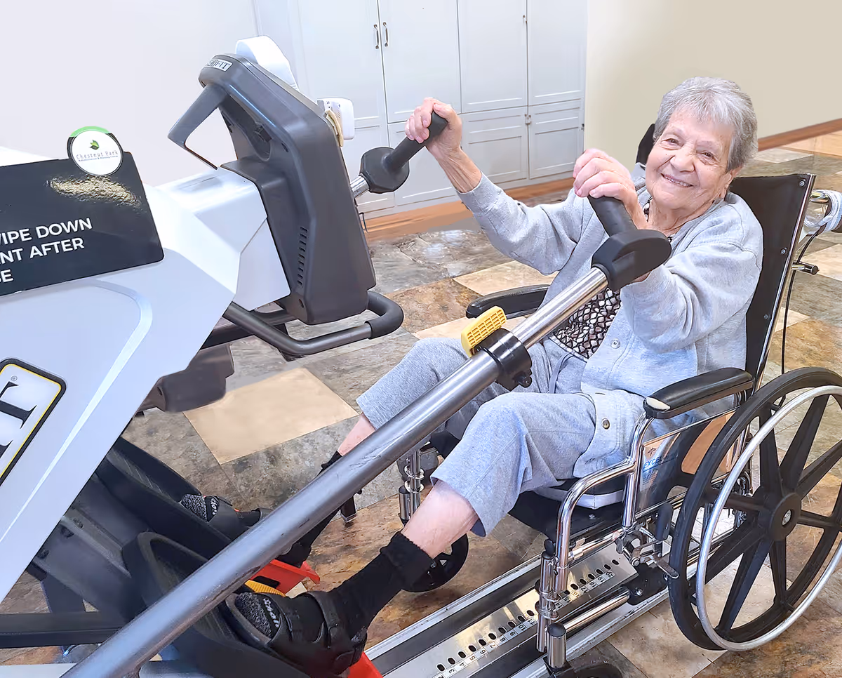 An elderly woman sitting in a wheelchair using a hand cycle exercise machine indoors, smiling at the camera. The room has a tiled floor and white cabinets in the background. A sign on the exercise machine reads 'Chestnut Park' and 'WIPE DOWN EQUIPMENT AFTER USE'.