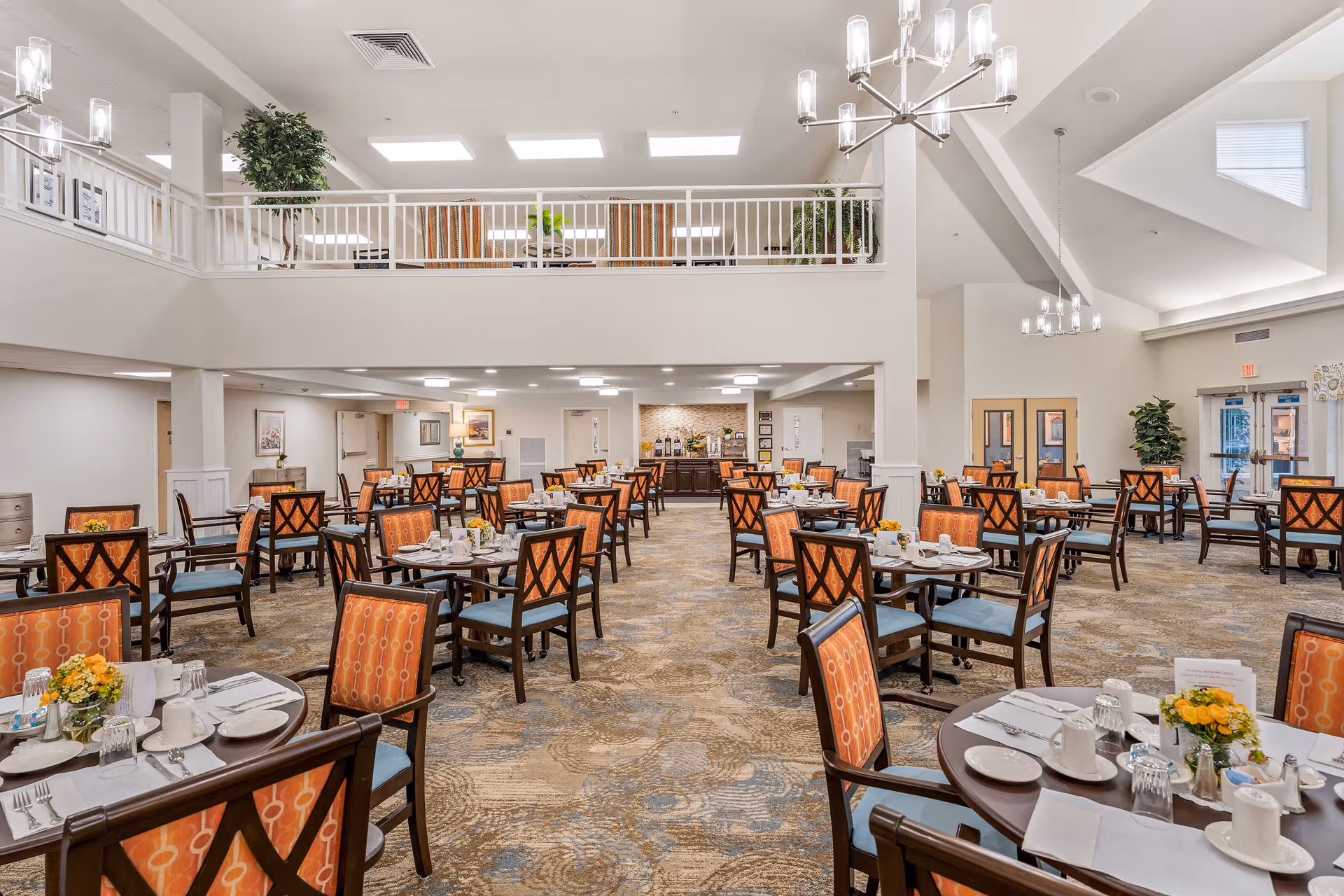 A spacious and well-lit dining room in a retirement living facility with multiple round tables set for meals. Each table has orange cushioned chairs with a patterned design, white tableware, and small floral centerpieces. The room features high ceilings with modern chandeliers, a carpeted floor with a patterned design, and a second-floor balcony with railings overlooking the dining area. There are plants and framed pictures on the walls, and a buffet area is visible at the far end of the room.