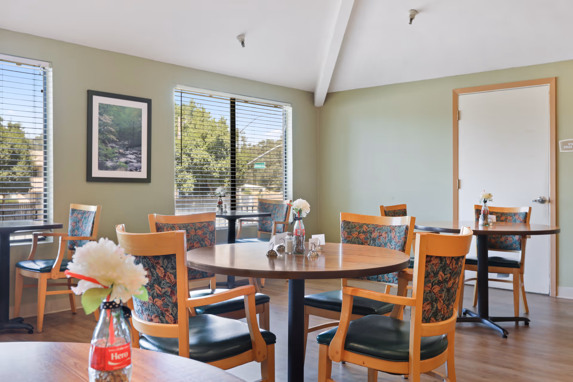 A bright dining room with round wooden tables and chairs featuring floral upholstery. Each table has a small vase with white flowers. Large windows with blinds allow natural light to fill the room, and a framed nature photograph hangs on the light green wall.