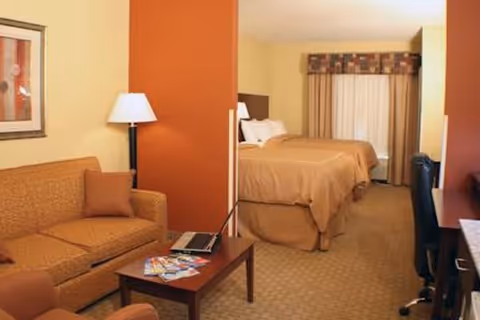 Interior view of a senior living facility room featuring a sitting area with a brown sofa, a coffee table with magazines and a laptop, a floor lamp, and two beds with beige bedding in the background. The room has warm-toned walls and carpet, a window with curtains, and a desk with a chair on the right side.