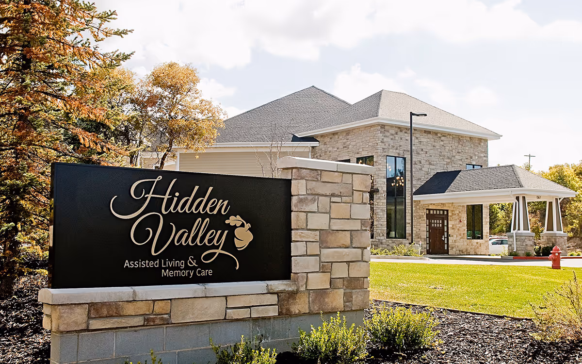 Exterior view of Hidden Valley Assisted Living and Memory Care facility with a stone building and a large sign in front displaying the facility's name. The area is landscaped with green grass, bushes, and trees with autumn foliage under a partly cloudy sky.