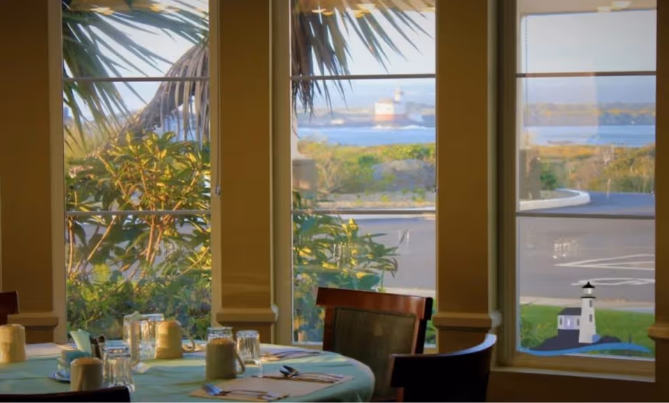 Dining area with a round table set with glasses, cups, and silverware. Large windows provide a view of greenery, a road, and a lighthouse in the distance.