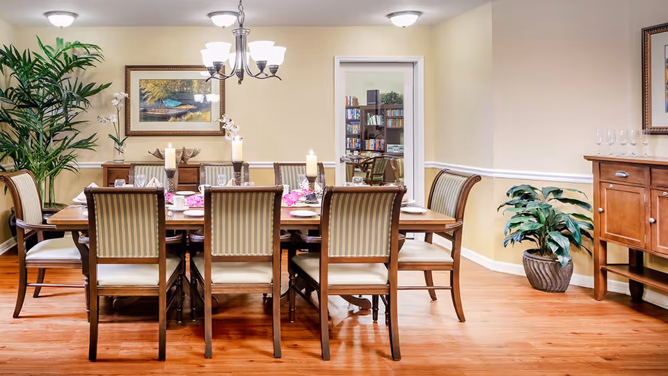 A well-lit dining room with a wooden dining table set for eight people with candles, plates, glasses, and pink flower decorations. The room has wooden flooring, beige walls, a chandelier, framed artwork, and two potted plants. A doorway in the back leads to another room with bookshelves and chairs.