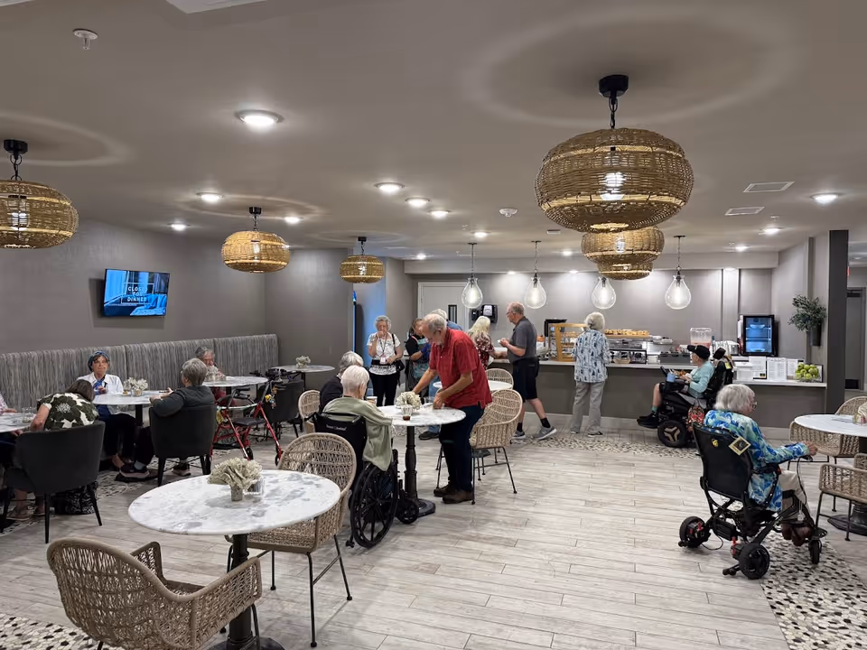 A dining area in a senior living facility with several elderly residents sitting at round tables and some standing near a buffet counter. The room has wicker pendant lights hanging from the ceiling, a TV mounted on the wall, and a modern, cozy atmosphere with light-colored flooring and neutral walls.