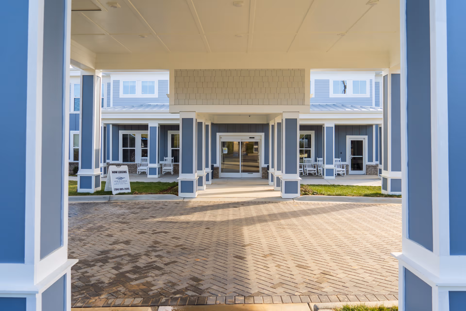Entrance of Arbor Ridge at Huntersville facility showing a covered driveway with blue and white pillars, a paved brick driveway, and a building facade with windows and rocking chairs on the porch. A sign near the entrance reads 'Now Leasing Arbor Ridge at Huntersville' with a phone number.