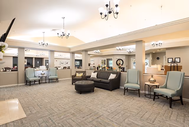 Spacious senior living common area with a sofa, armchairs, round ottoman, chandeliers, and a reception desk in the background.