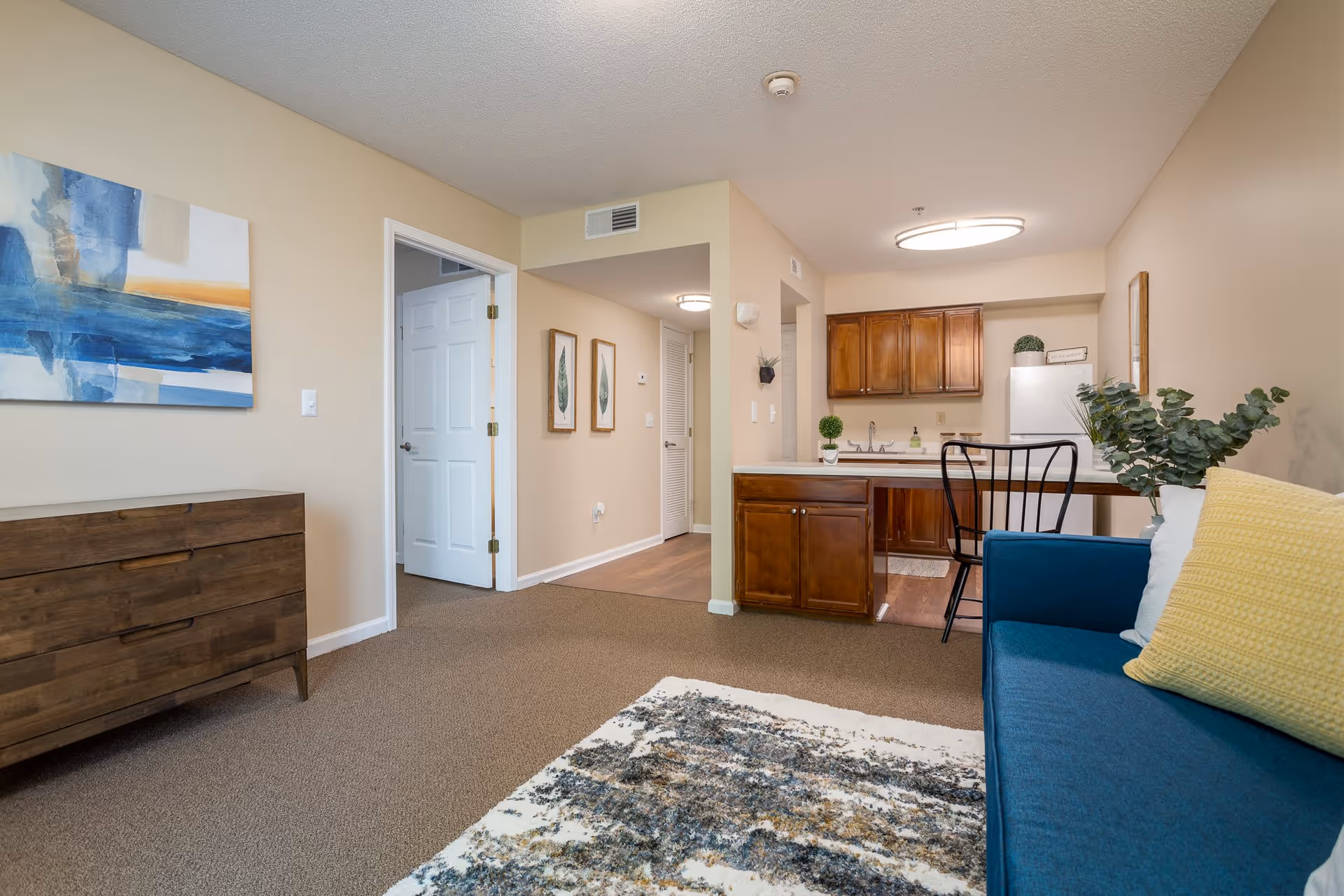 Interior view of a senior living facility apartment showing a living area with a blue couch with yellow and white pillows, a patterned rug, a wooden dresser, and a small kitchen with wooden cabinets, a white refrigerator, and a counter with a black chair. The walls are beige with some framed artwork, and the floor is carpeted with a transition to wood in the kitchen area.