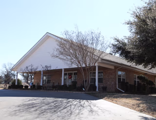 Front exterior of a single-story brick building with a covered entrance, columns, and surrounding trees.