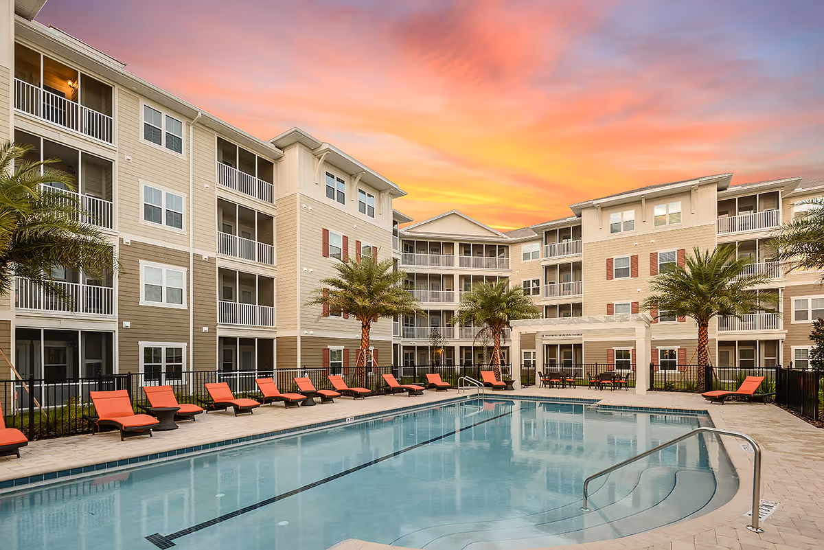 Outdoor swimming pool with lounge chairs in front of a multi-story apartment building at sunset.