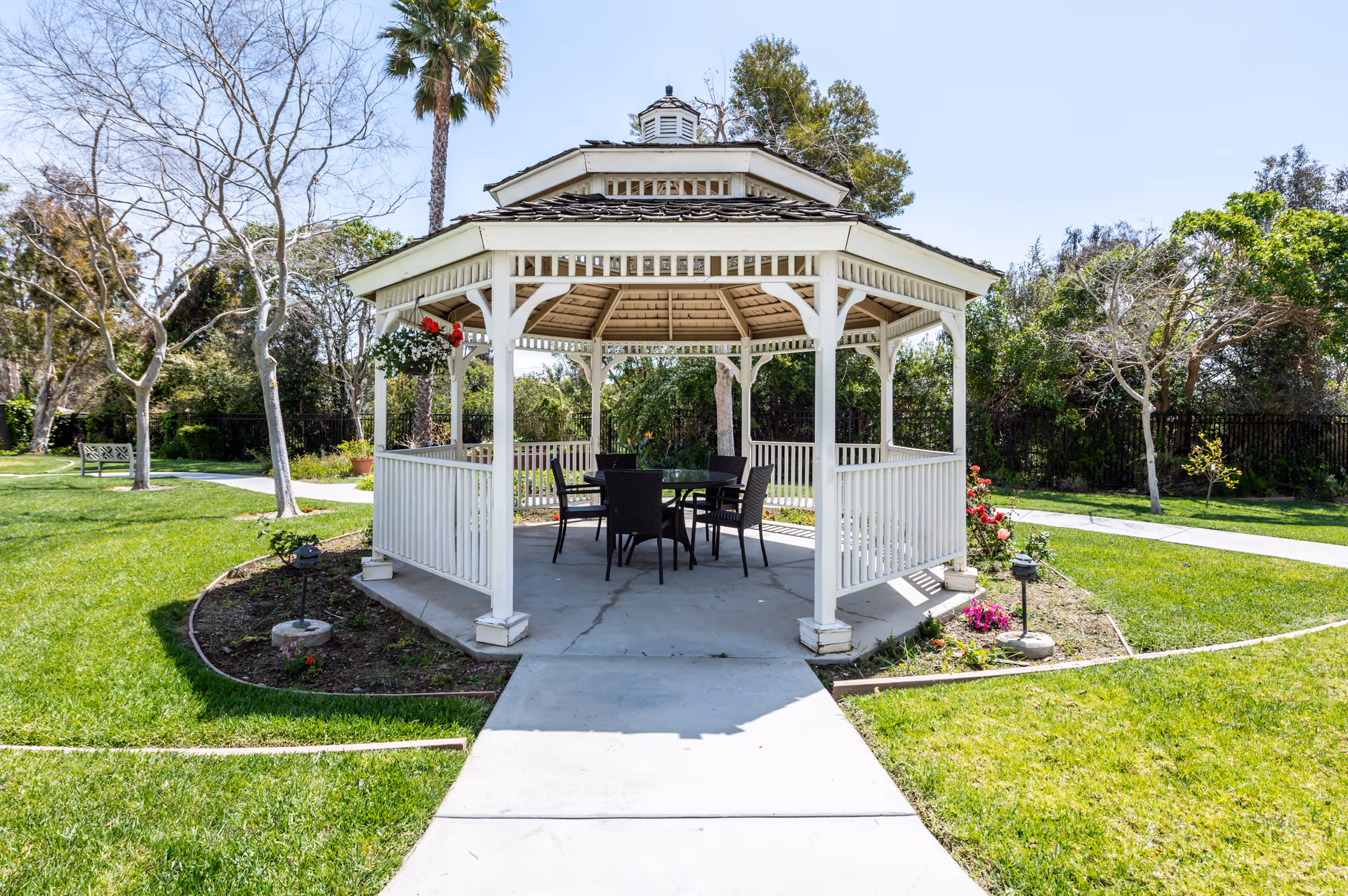 A white wooden gazebo with a shingled roof situated in a green garden area. Inside the gazebo, there is a round glass table surrounded by four black chairs. The gazebo is surrounded by grass, flower beds, and trees, with a concrete pathway leading up to it under a clear blue sky.