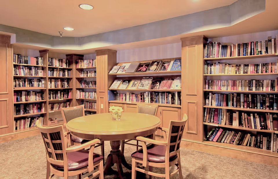 A cozy library room with wooden bookshelves filled with books along the walls. In the center, there is a round wooden table surrounded by four wooden chairs with cushioned seats. A small vase with flowers is placed on the table. The room has soft lighting from ceiling fixtures and a carpeted floor.