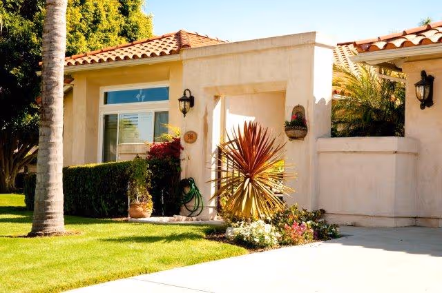 Exterior view of a single-story residential building with beige stucco walls and a red tile roof. The entrance is framed by a small garden with various plants and flowers, including a tall palm tree and a spiky red-leafed plant. There is a wall-mounted lantern light near the doorway and a garden hose coiled on the wall.