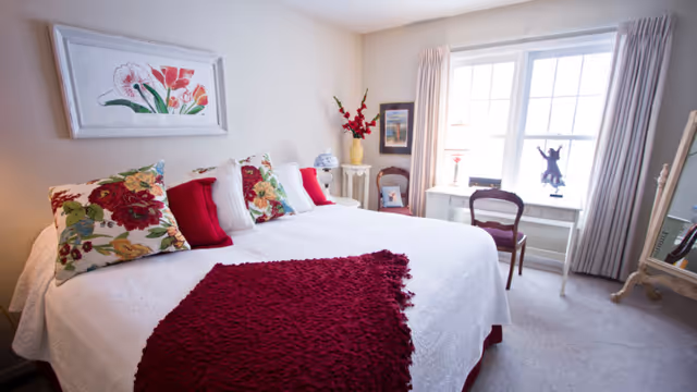 Bright bedroom with a large bed dressed in white linens and red accents, a desk by a window, and floral artwork on the wall.