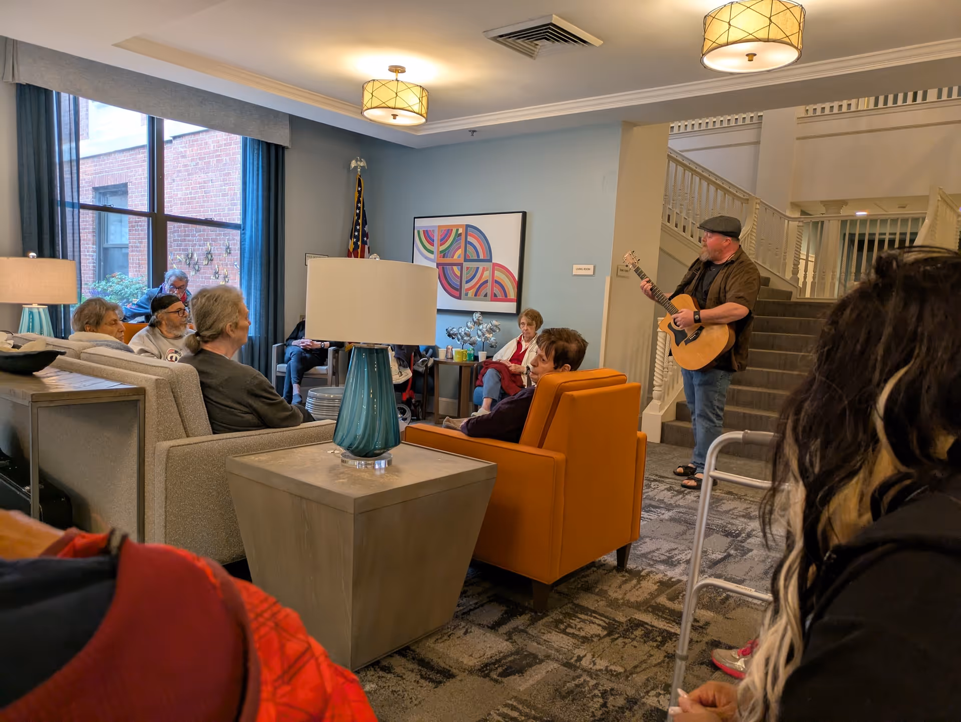 A group of elderly people seated in a living room area of a senior living facility, attentively listening to a man playing an acoustic guitar near a staircase. The room has comfortable chairs, a large window with blue curtains, modern lamps, and colorful abstract artwork on the wall.