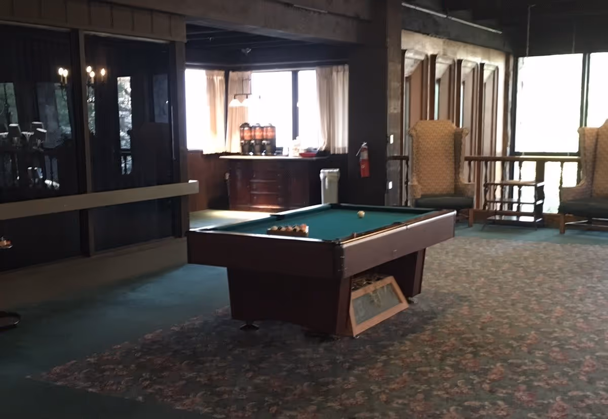 Interior room with a pool table in the center, two upholstered armchairs near large windows, and a cabinet with beverage dispensers in the background. The room has carpeted flooring and wooden paneling on the walls.
