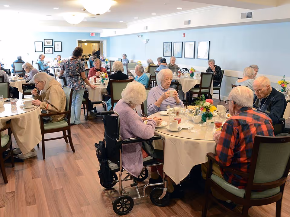 A dining room in a senior living facility with elderly residents seated at round tables covered with white tablecloths, eating and drinking. A staff member is standing and assisting one of the tables. The room has wooden flooring, light blue walls with framed pictures, and ceiling lights.