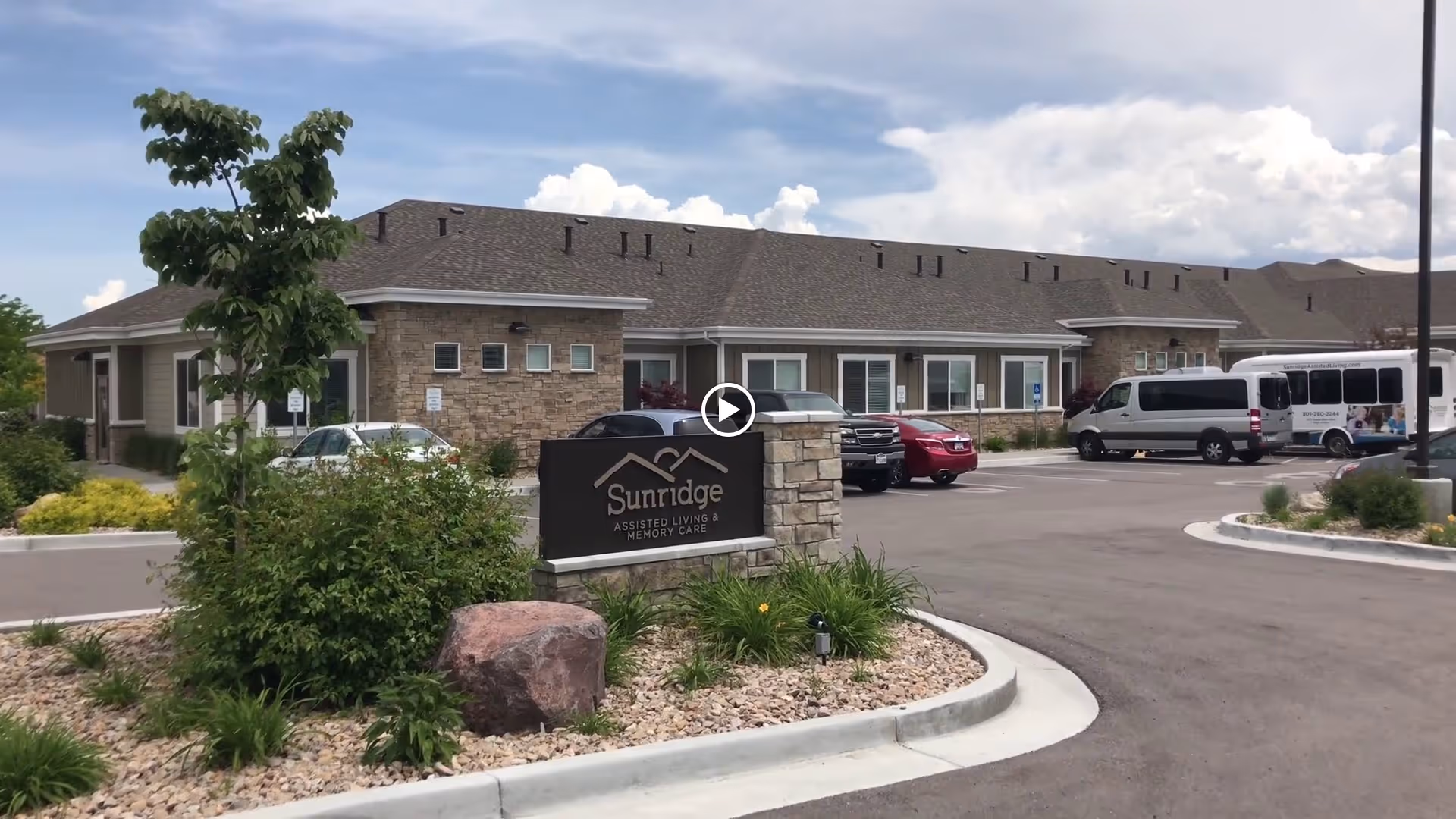 Exterior view of Sunridge Assisted Living and Memory Care facility showing a single-story building with stone and siding facade, a parking lot with several vehicles including vans and cars, landscaped greenery with bushes, a tree, and rocks, under a partly cloudy sky.