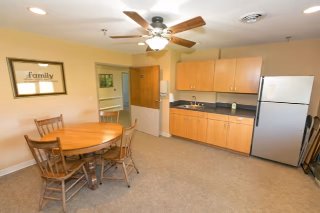 A room with a round wooden table and four wooden chairs, a ceiling fan with lights, wooden cabinets with a sink, and a stainless steel refrigerator. There is a framed picture on the wall and an open door leading to another room.