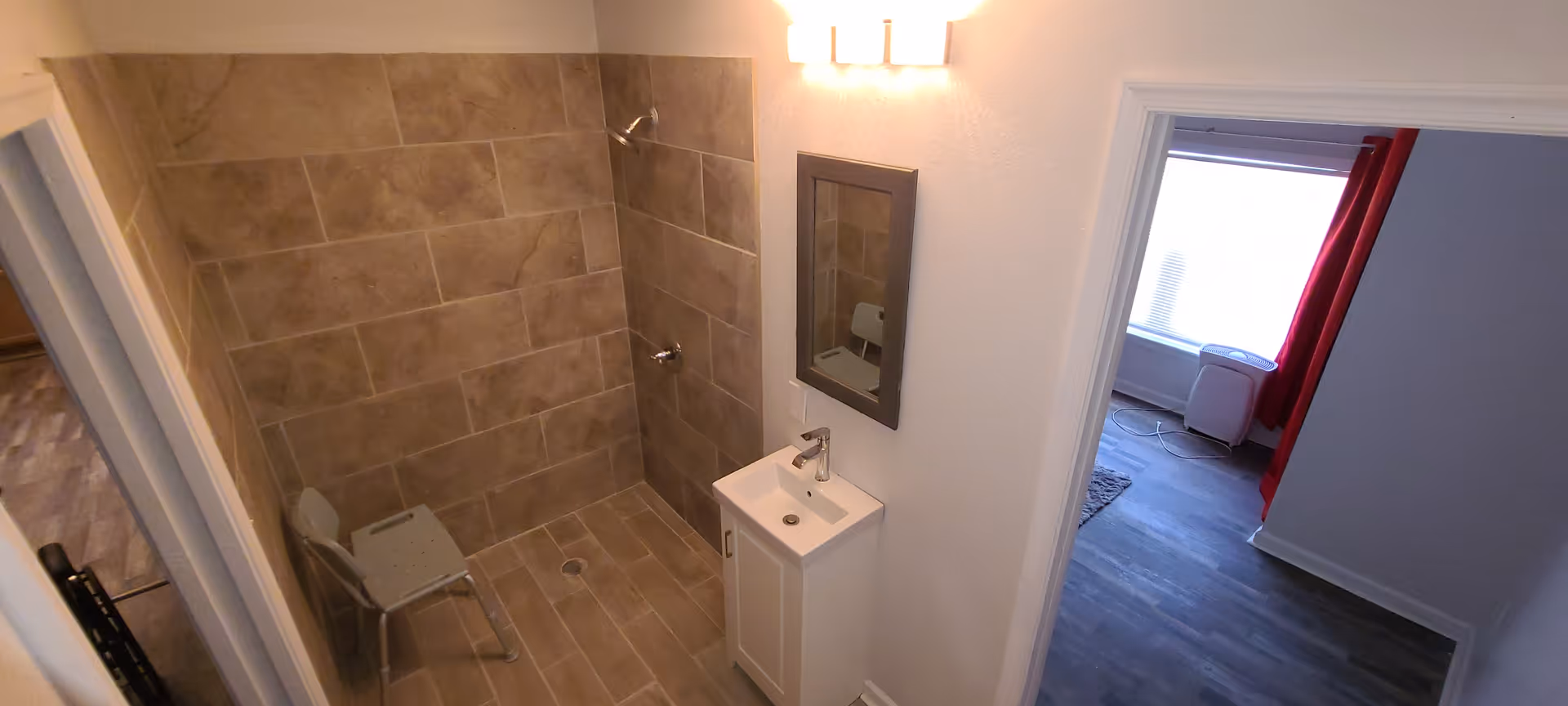 A small bathroom with beige tiled walls and floor, featuring a shower area with a metal chair, a white sink with a cabinet, a mirror above the sink, and a light fixture above the mirror. Adjacent to the bathroom is a room with dark wood flooring, a window with red curtains, and a white air purifier or heater.