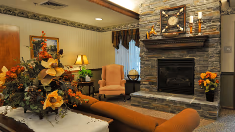 A cozy living room area featuring a stone fireplace with a clock and decorative items on the mantel. There is a pink upholstered armchair next to a small table with a lamp and a globe. A brown sofa is partially visible in the foreground, and a large floral arrangement is on a white table. The room has warm lighting and traditional decor.