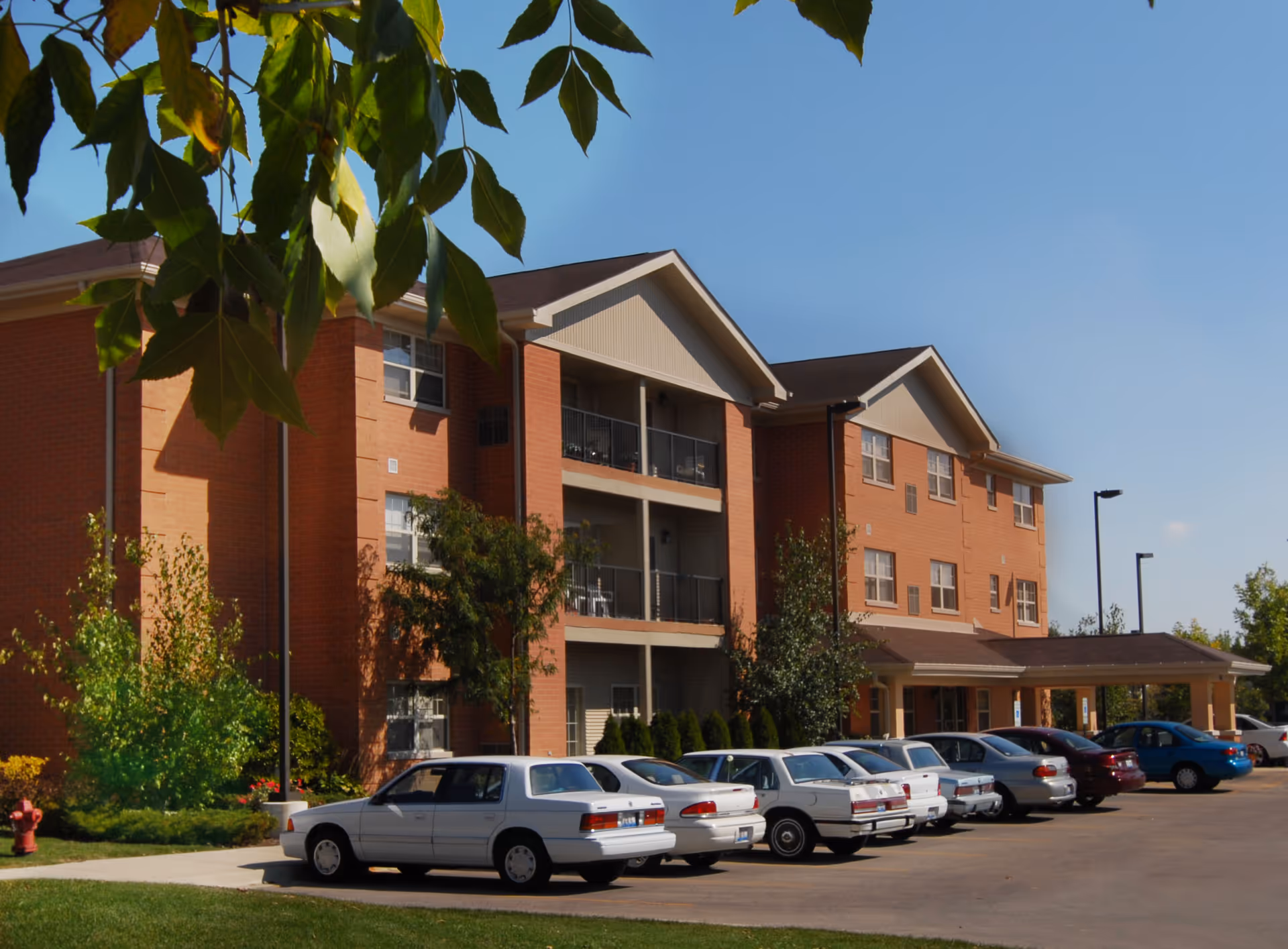 Exterior view of a three-story brick senior living building with balconies and a row of parked cars in front under a clear sky.