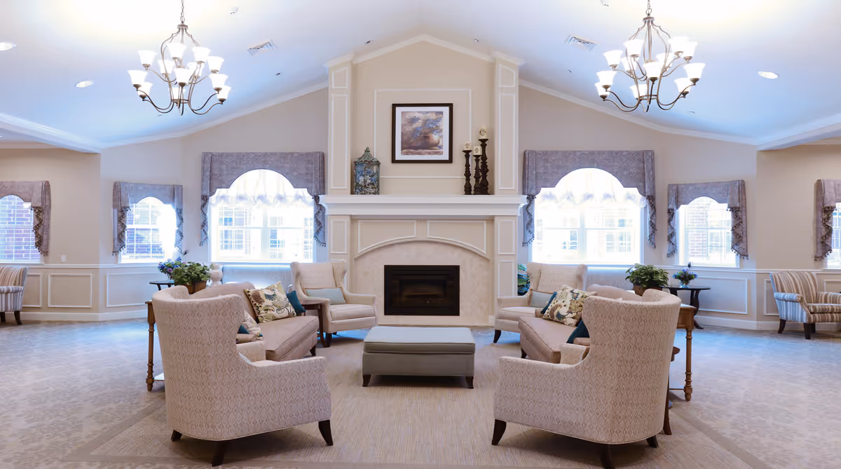 A spacious and well-lit living room with a high vaulted ceiling, two chandeliers, and large windows with decorative curtains. The room features a central fireplace with a framed picture and decorative items on the mantel. Comfortable upholstered armchairs and sofas are arranged around a central ottoman, with side tables and potted plants adding to the cozy atmosphere.