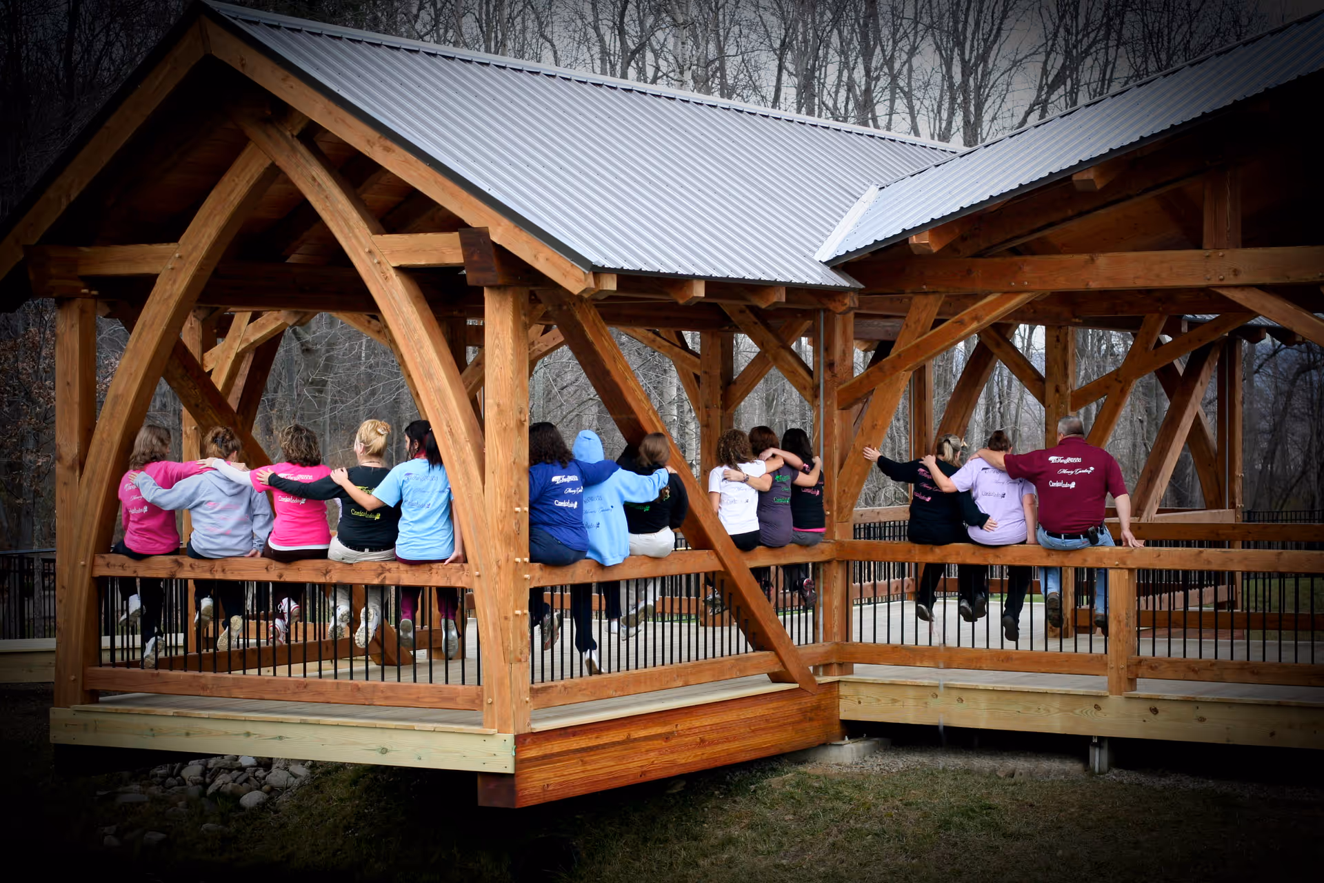 A group of people sitting closely together on a wooden railing under a covered wooden pavilion, with their arms around each other, facing away from the camera. The pavilion is set in an outdoor area with leafless trees in the background.