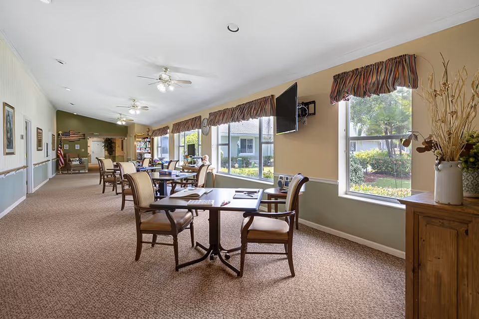 A bright and spacious common area in a senior living facility with several tables and chairs arranged along large windows that let in natural light. The room has beige walls with a green lower half, carpeted floors, ceiling fans with lights, and decorative window valances. There is a wall-mounted TV and a wooden cabinet with a vase of dried plants. Outside the windows, greenery and part of the building exterior are visible.