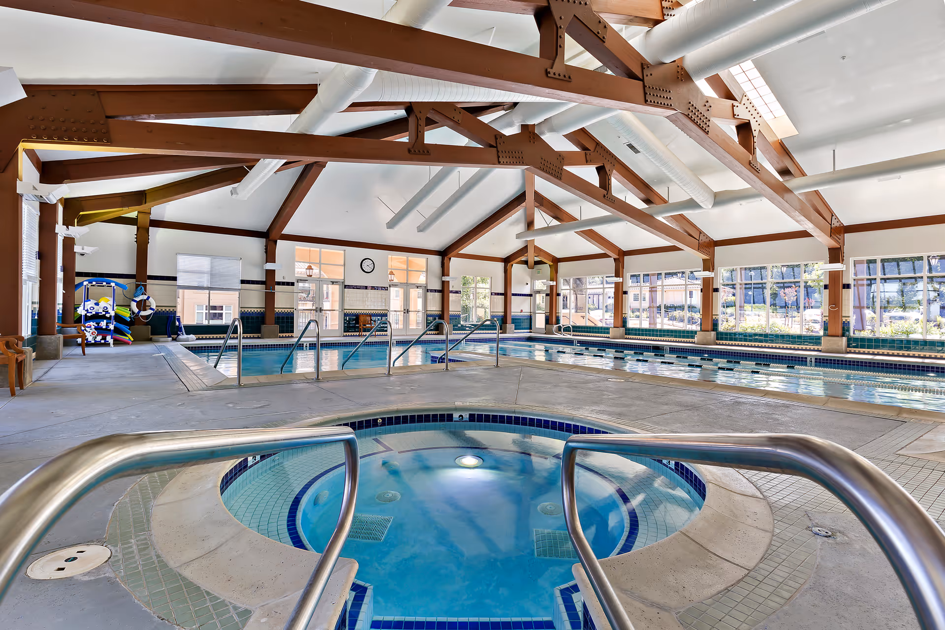 Indoor swimming pool area with a hot tub in the foreground, large windows letting in natural light, and exposed wooden beams on the ceiling.