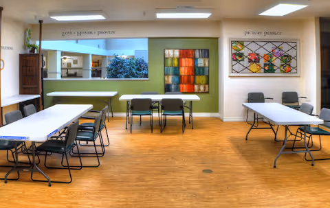 A spacious room with several rectangular tables and chairs arranged around them, featuring wooden flooring and light-colored walls. The back wall has two colorful artworks and a large open window-like cutout showing greenery outside. The room is well-lit with ceiling lights and has motivational words on the walls such as 'LOVE • HONOR • PROVIDE' and 'PICTURE PERFECT'.