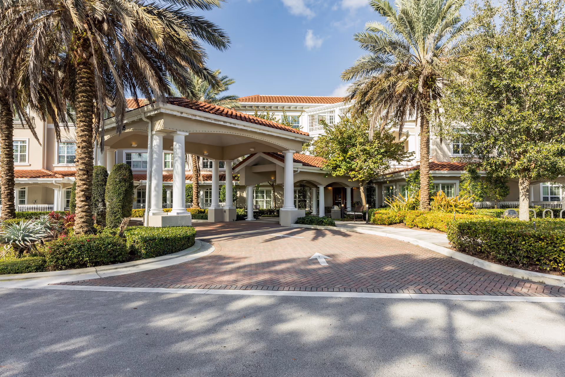 Entrance to a senior living facility with a covered driveway supported by white columns, surrounded by palm trees and well-maintained landscaping under a blue sky.
