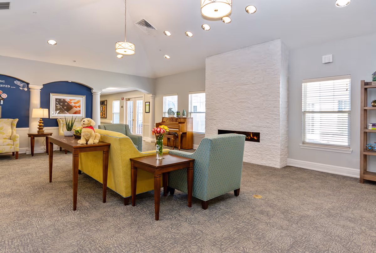 A bright and spacious living room area in Dorset Place Memory Care featuring comfortable seating including a yellow sofa and blue armchairs arranged around wooden tables. A small stuffed dog toy sits on one table, and a vase with flowers is on another. The room has a modern white fireplace, a piano near the windows, and soft lighting from ceiling fixtures. The carpet has a subtle patterned design, and the walls are painted light gray with white trim.