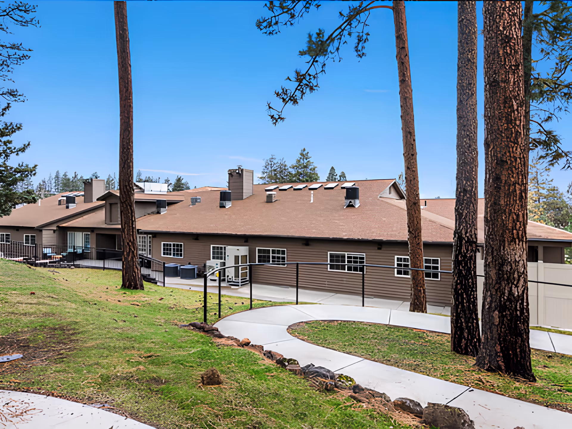 Brown single-story memory care building with a curved concrete walkway and tall pine trees in the foreground.